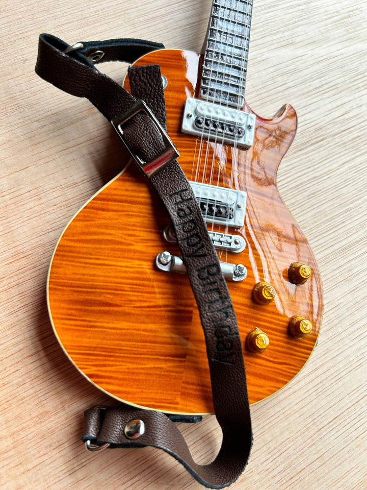 A dark tan leather miniature guitar strap fitted on a miniature guitar with a brown body and cream pickguard, displayed on a wooden surface.