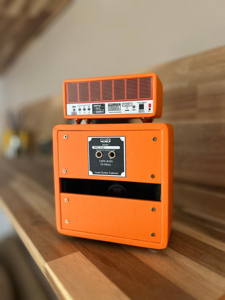 A miniature orange-colored Orange brand amplifier, featuring a speaker cabinet and a control panel with knobs and branding, placed on a wooden surface.