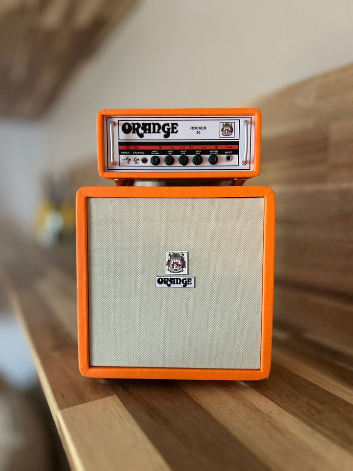 A handcrafted miniature orange-colored Orange brand amplifier, featuring a speaker cabinet and a control panel with knobs and branding, placed on a wooden surface.