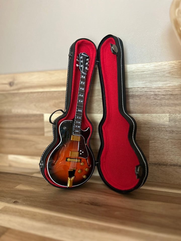 Red miniature guitar case with black trim open on a wooden shelf with electric guitar inside