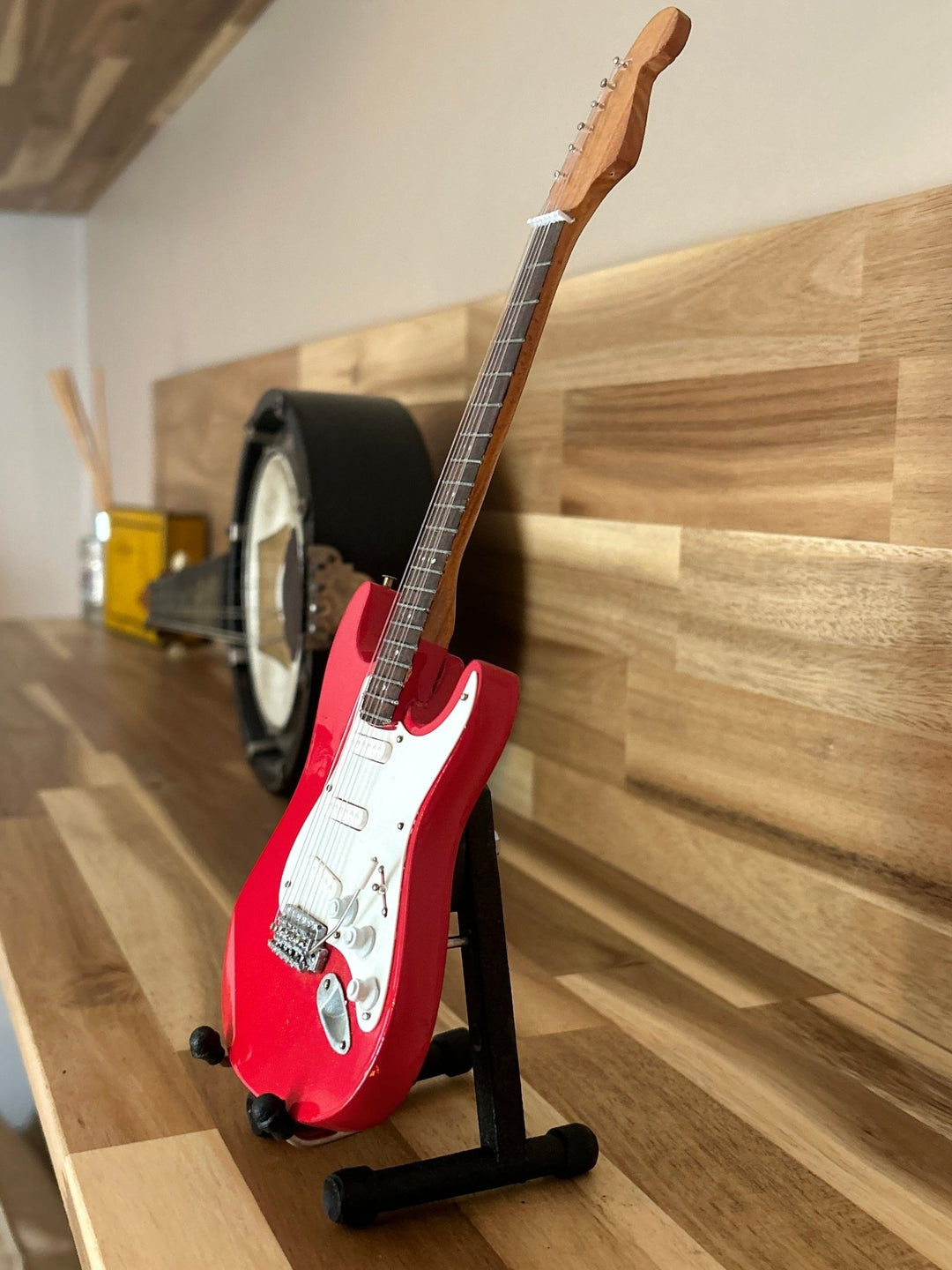 Red electric miniature Stratocaster guitar on a stand against a wooden wall.