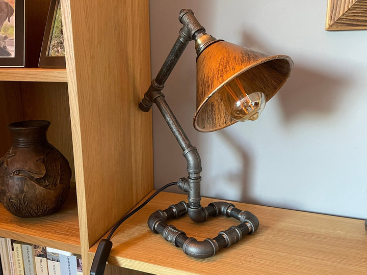 Industrial-style desk lamp with copper coloures shade on a wooden surface next to a bookshelf.